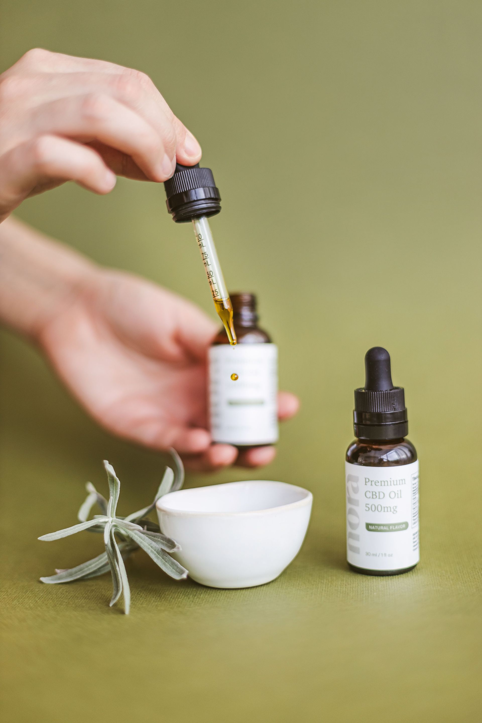 Close-up of hands holding a CBD oil dropper above a white bowl on a green background.